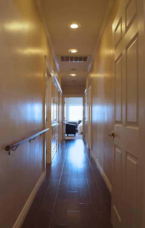 A narrow hallway with wooden flooring and beige walls, featuring a handrail on the left side and several closed doors on both sides. Recessed ceiling lights illuminate the corridor, and at the end of the hallway, a room with a window, a chair, and a bed is visible.
