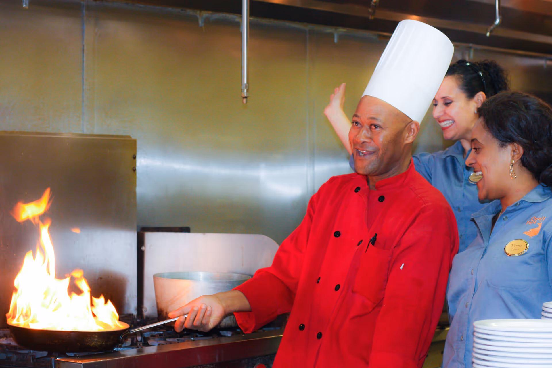 A chef in a red uniform and tall white chef hat is cooking with a flaming pan on a stove in a commercial kitchen. Two women in blue uniforms stand beside him, smiling and watching the cooking process.