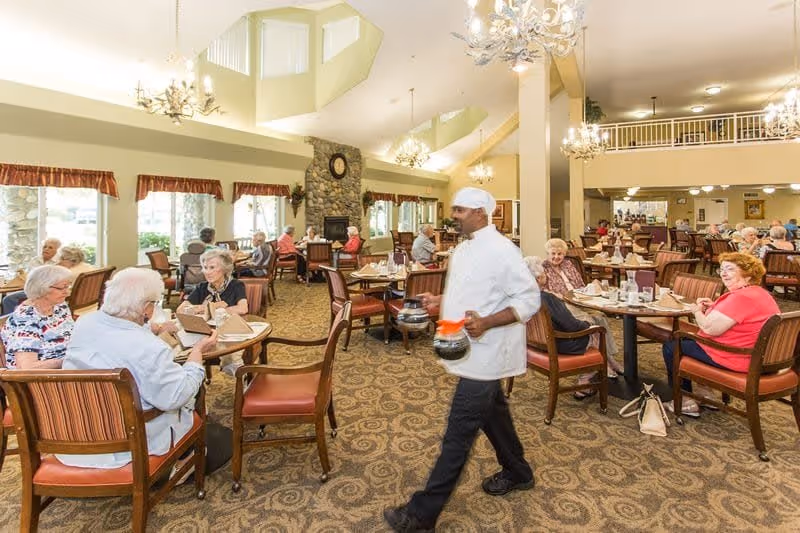 A spacious dining room in Creekside Oaks Retirement Community with elderly residents seated at tables, enjoying meals and conversation. A staff member dressed in a white uniform and hat is walking through the room carrying coffee pots. The room features large windows, a stone fireplace with a clock above it, chandeliers, and a patterned carpet.