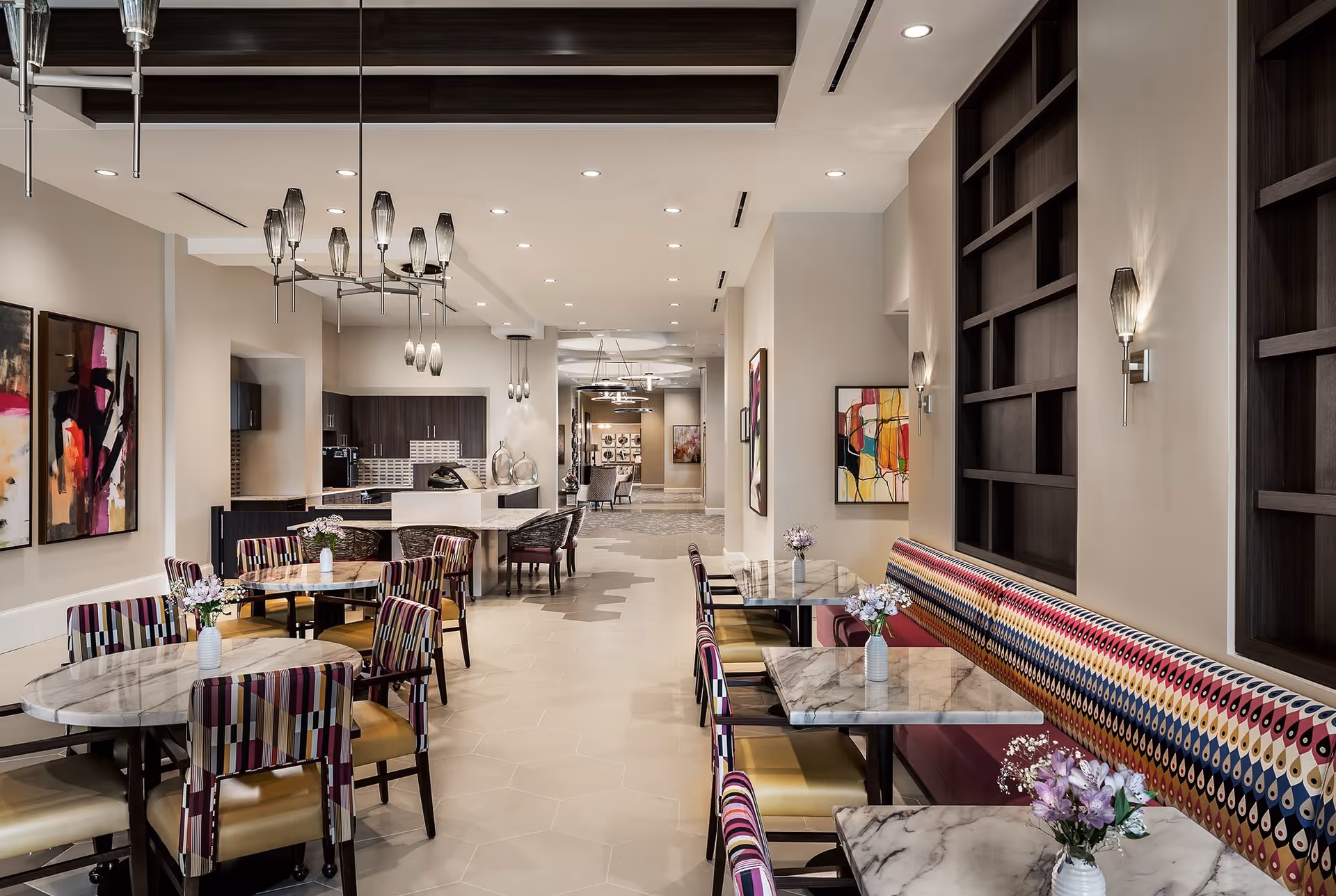 A modern dining area in a senior living facility featuring marble-top tables with colorful patterned chairs and a long bench with a vibrant geometric design. The room is well-lit with contemporary pendant lights and wall sconces, and decorated with abstract artwork on the walls. In the background, there is a kitchen area with dark wood cabinetry and a countertop.