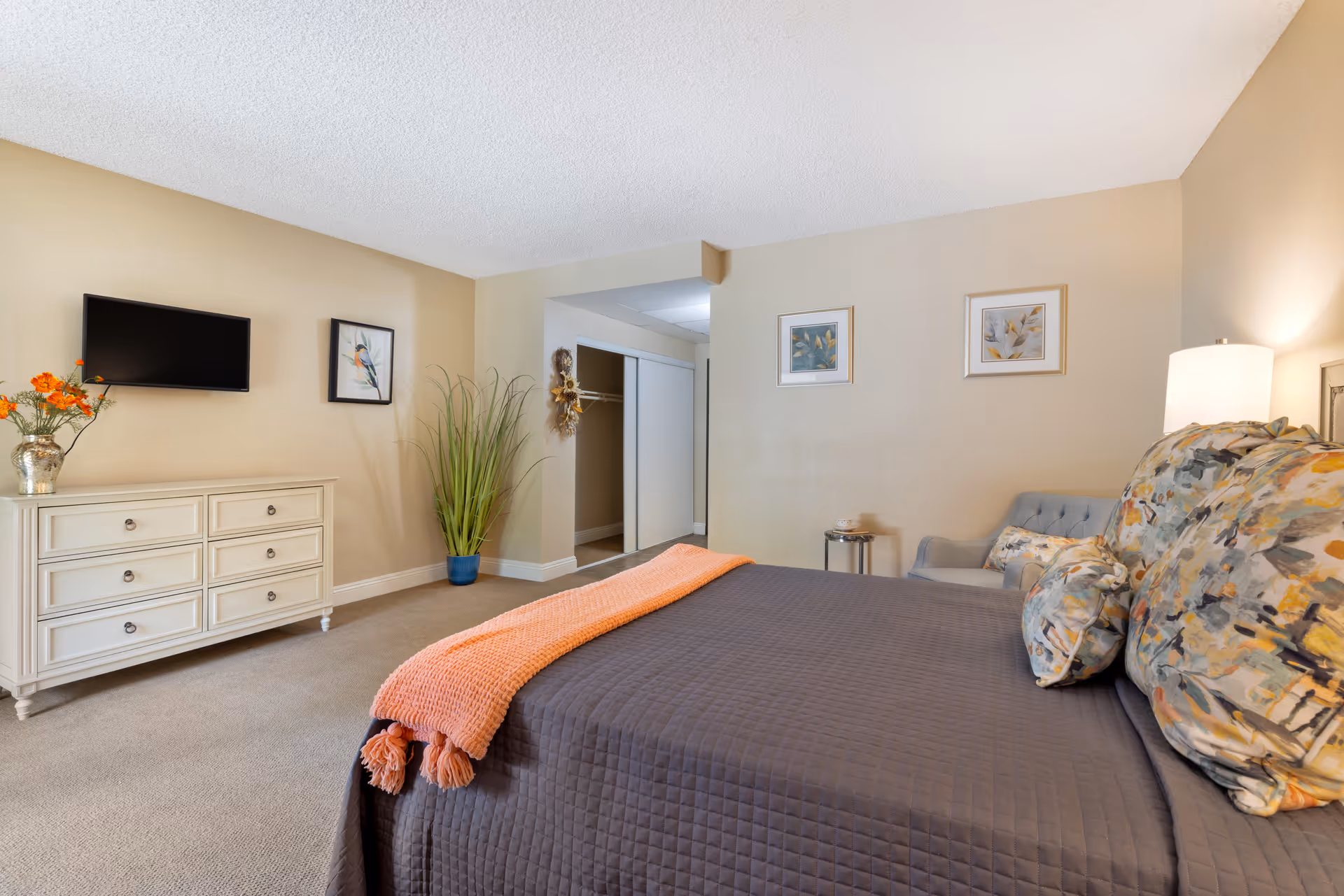 A cozy bedroom with a large bed covered in a dark quilt and floral pillows, an orange throw blanket at the foot of the bed, a white dresser with a vase of orange flowers, a wall-mounted TV, a potted plant, an open closet with sliding doors, and framed artwork on beige walls.
