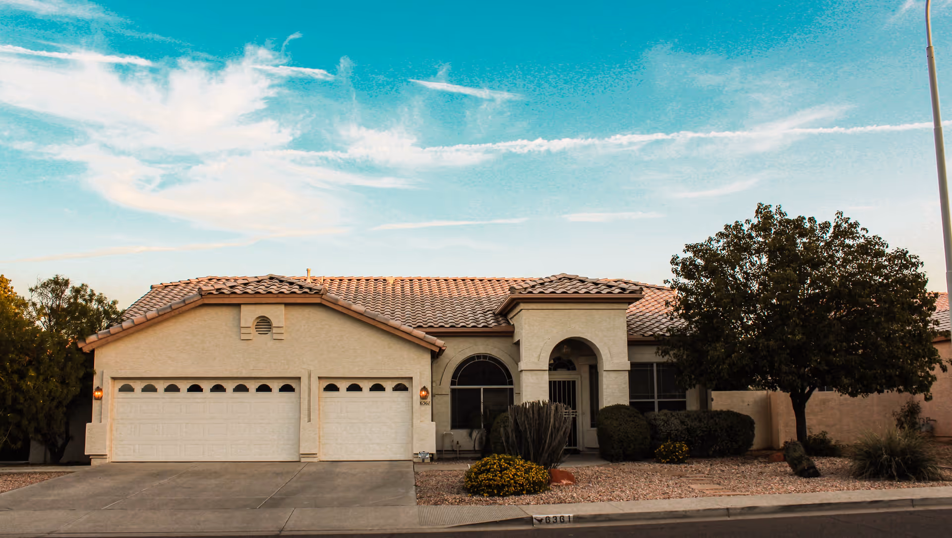 Exterior view of a single-story residential building with a tiled roof, three garage doors, and a front entrance with an archway. The front yard is landscaped with gravel, bushes, a tree, and some yellow flowers. The sky is clear with some clouds.