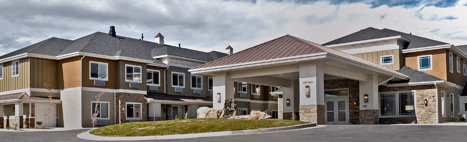 Exterior view of a senior living facility building with a covered entrance, multiple windows, and a mix of stone and siding on the facade under a cloudy sky.