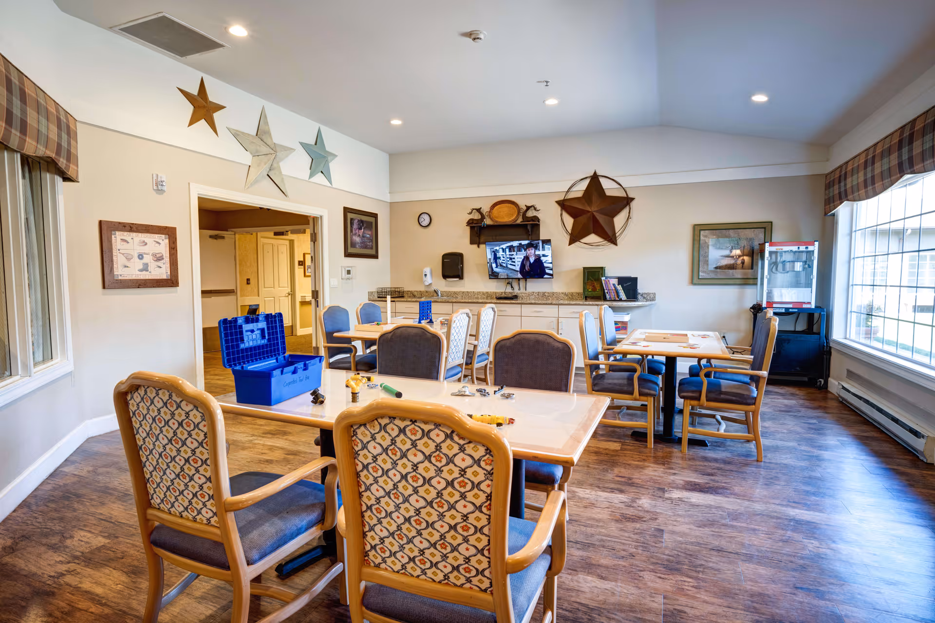 A bright and spacious activity room with multiple tables and chairs arranged for group activities. The room features large windows with plaid valances, wooden flooring, and walls decorated with large star ornaments and framed pictures. A television is mounted on the far wall above a counter with cabinets, and a popcorn machine is visible in the corner.
