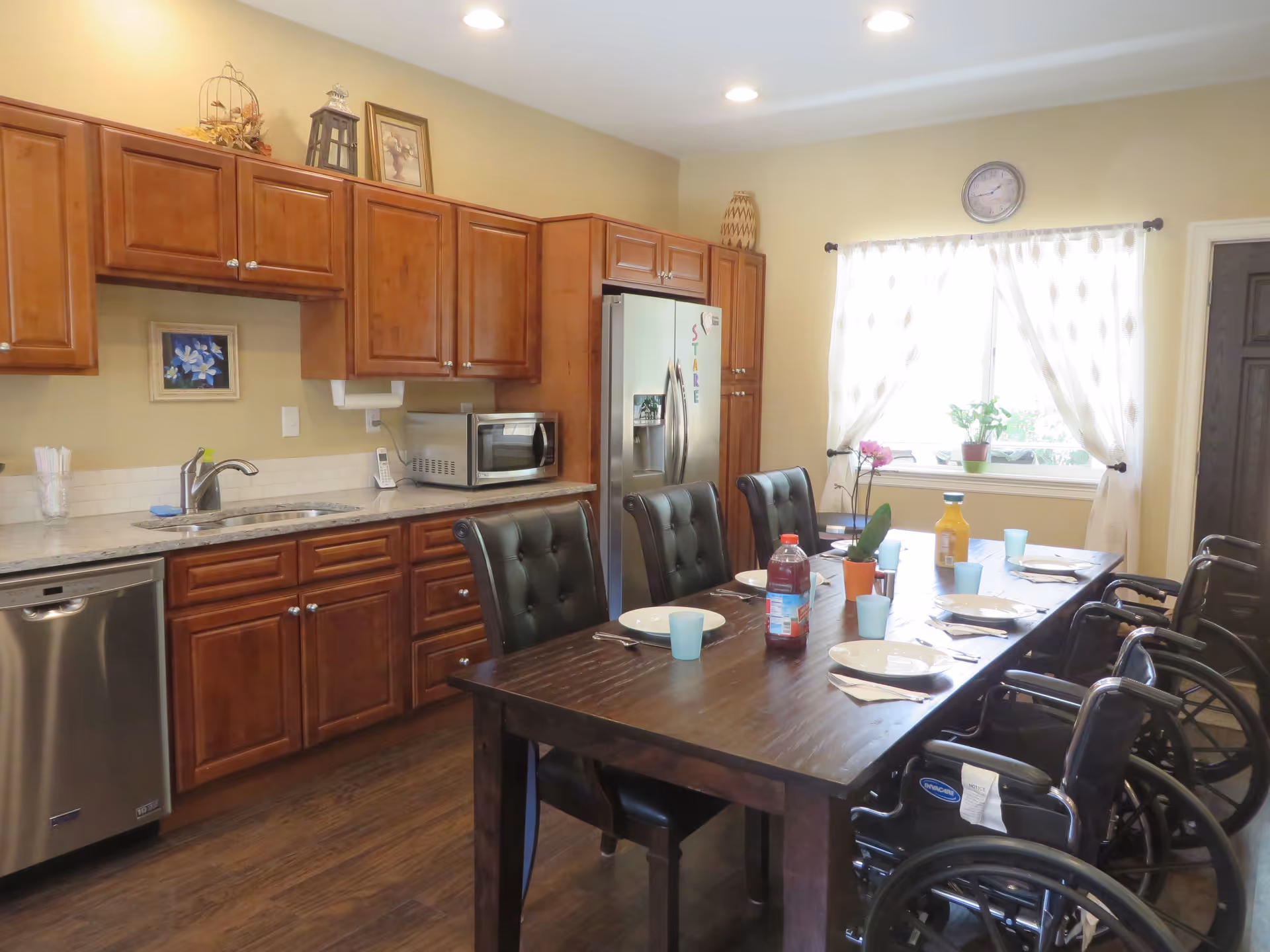 Dining room with a long table, wheelchairs, and wooden kitchen cabinets along the wall.