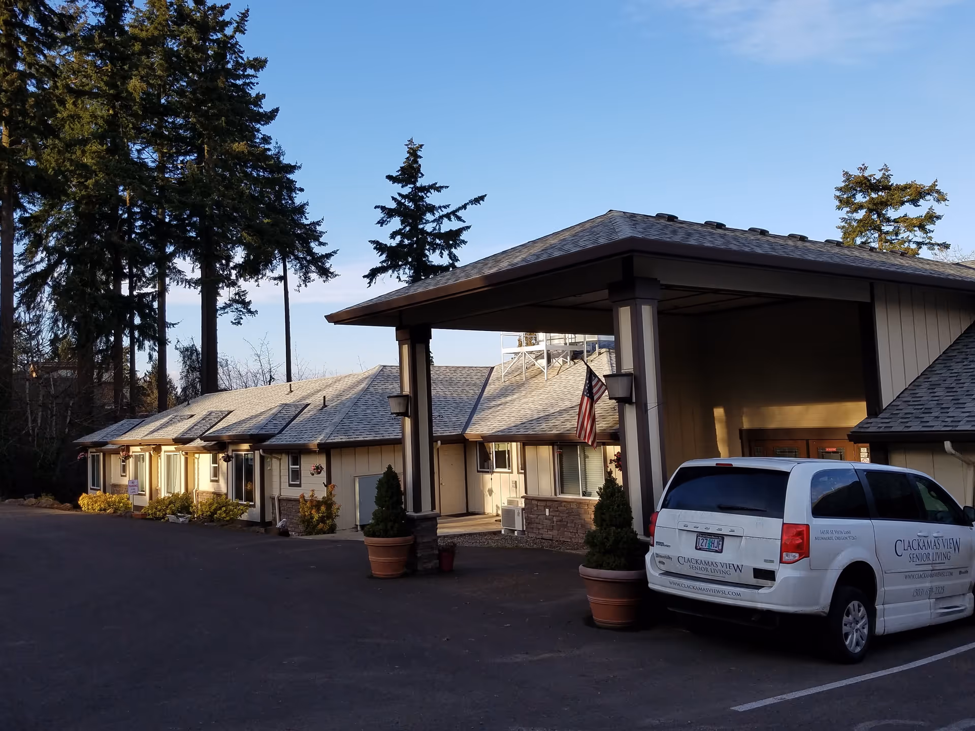Entrance and front facade of a senior living building with a covered drop-off, a parked service van, potted plants, and tall trees behind.
