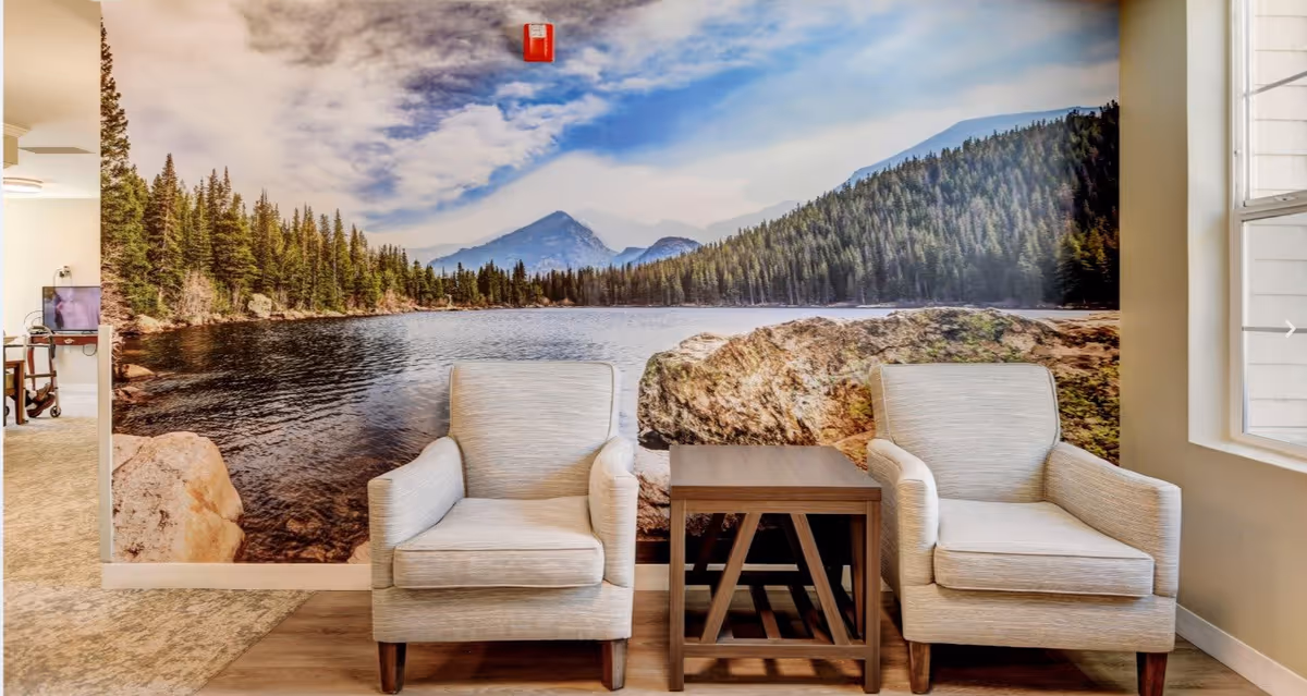 Two beige armchairs with wooden legs are positioned on either side of a small wooden table in a room. Behind them is a large wall mural depicting a scenic lake surrounded by pine trees and mountains under a partly cloudy sky. A window is visible on the right side, allowing natural light into the space.