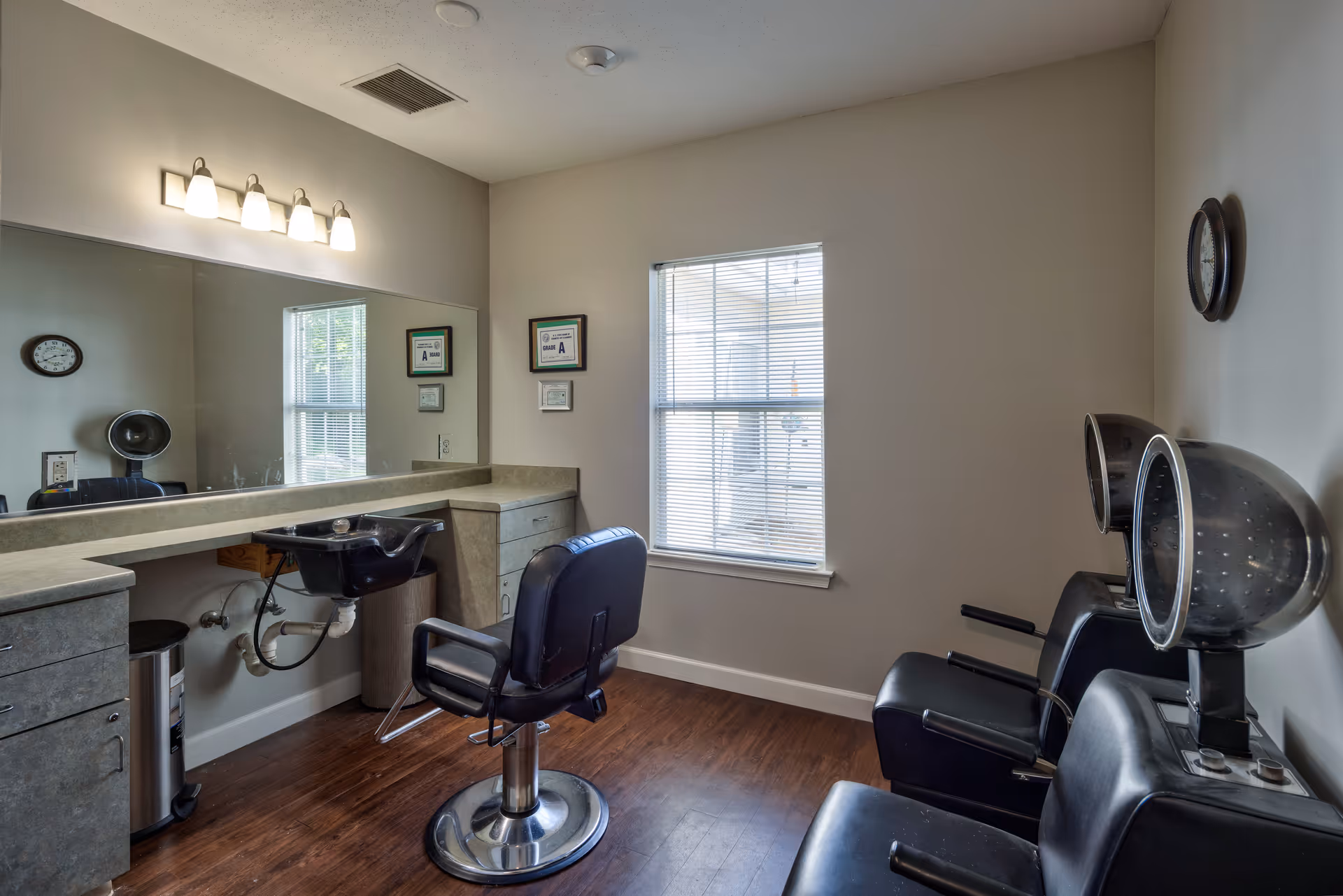 Interior of a salon room with a black salon chair in front of a sink and a large mirror. There are two black hair drying chairs with hooded dryers on the right side. The room has wooden flooring, a window with blinds, a clock on the wall, and certificates hanging near the window.