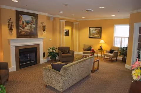 A cozy communal living room with a fireplace, sofa and armchairs under warm yellow walls and framed artwork.