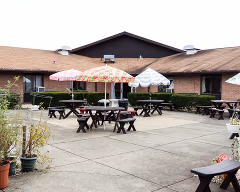 Outdoor courtyard area of a senior living facility with multiple picnic tables and benches. Several tables have colorful umbrellas providing shade. The building surrounding the courtyard is single-story with a brown roof and brick walls. Potted plants and shrubs are placed around the area.