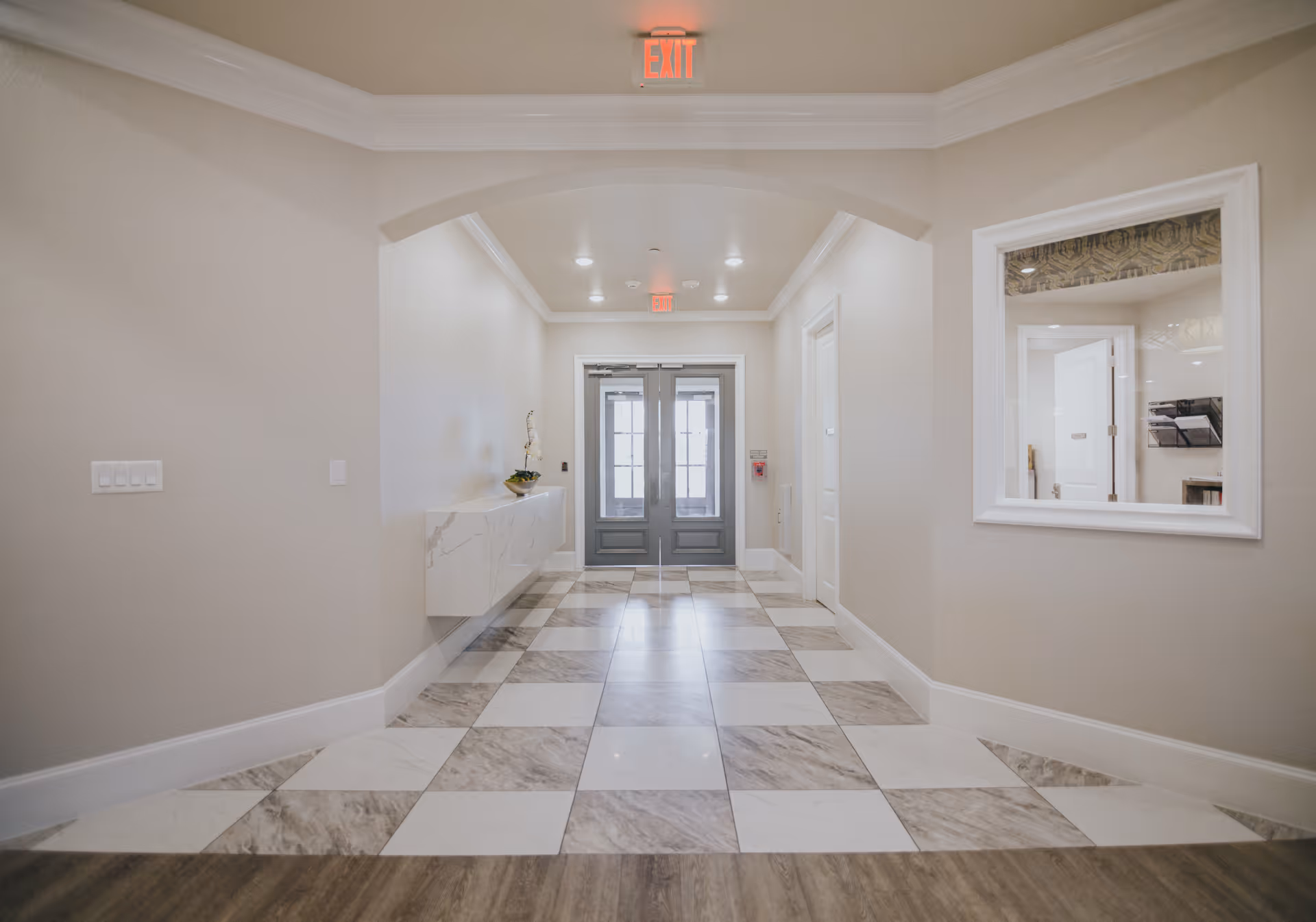 A clean, well-lit hallway with checkered tile flooring leading to double glass doors. The walls are painted light beige, and there is a floating marble console table with a small plant on the left side. A large window on the right wall looks into another room. An exit sign is visible above the doors.