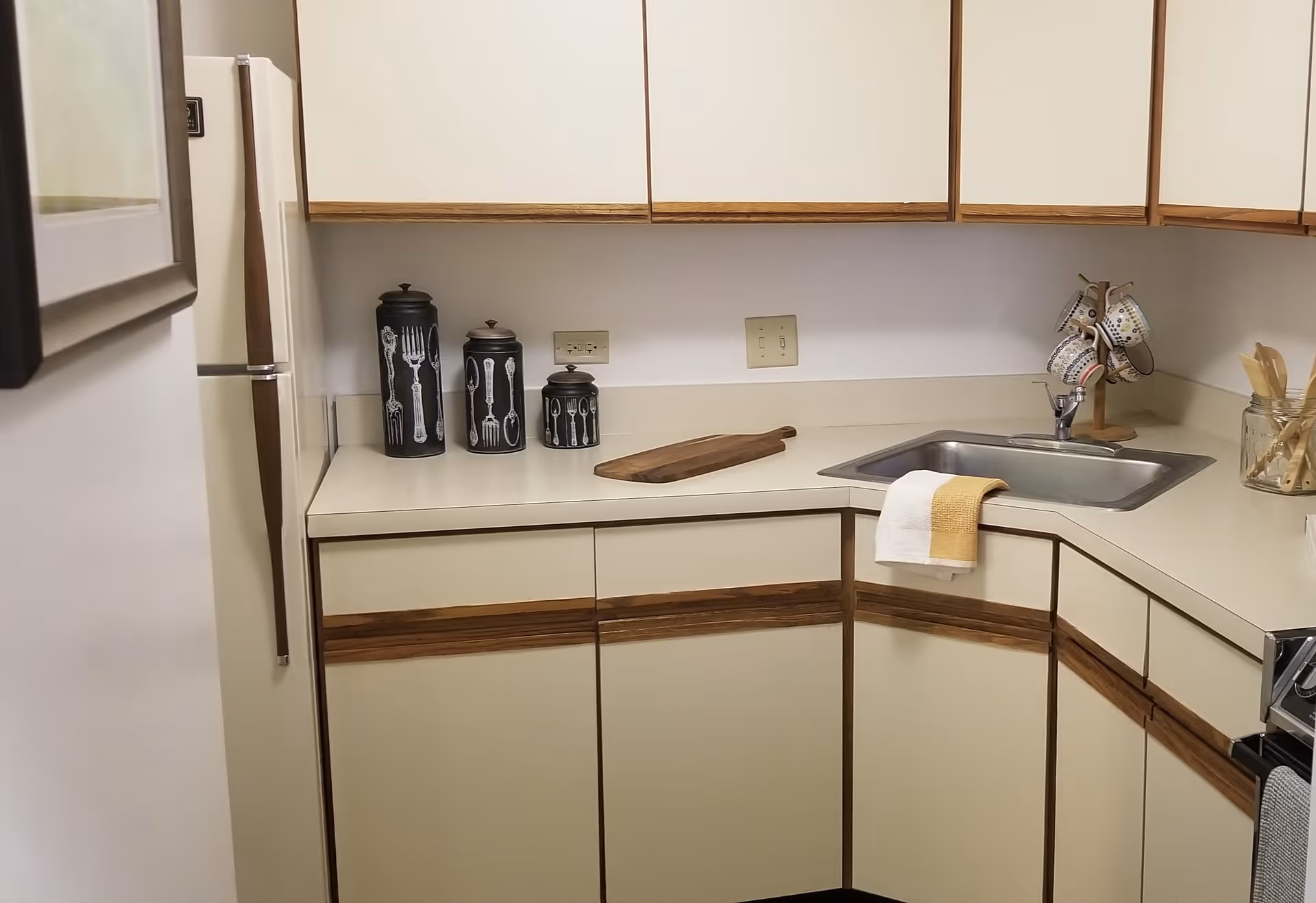L-shaped kitchen with cream cabinets, a countertop sink, refrigerator, decorative canisters and a mug rack.