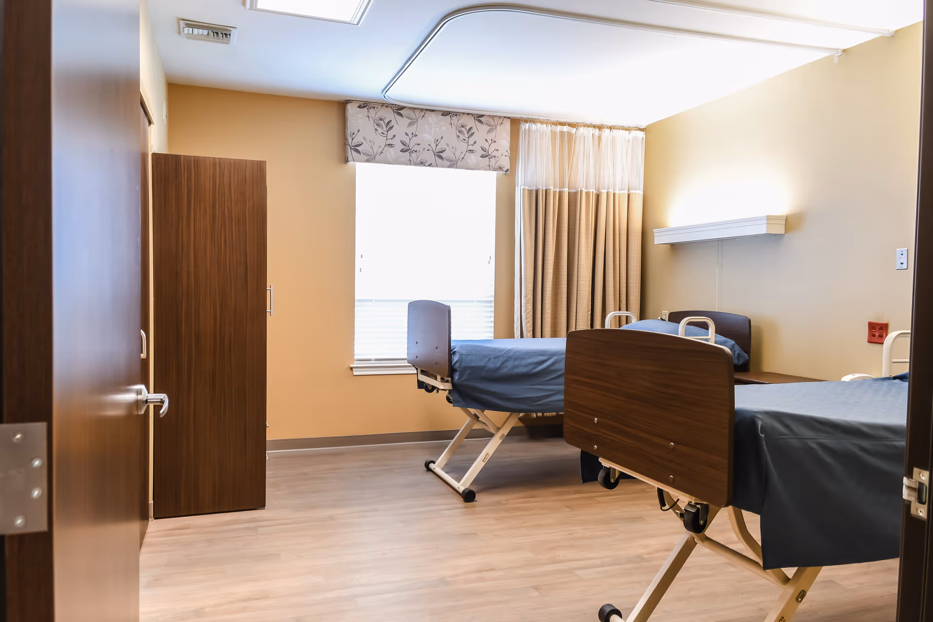 A room with two hospital-style beds with dark wood headboards and footboards, each covered with blue bedding. The room has light-colored walls, a window with floral and beige curtains, a wooden wardrobe, and a light wood floor. The door to the room is open, showing a silver door handle.