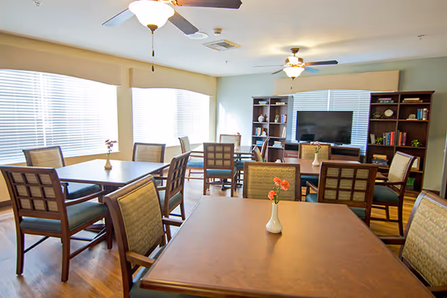 A bright and spacious common area with multiple wooden tables and cushioned chairs arranged neatly. Each table has a small vase with flowers. The room features large windows with blinds, ceiling fans with lights, and a large flat-screen TV mounted on a wooden shelving unit filled with books and decorative items.