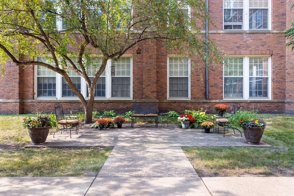 Outdoor seating area with metal chairs and a bench arranged on a concrete patio under a tree, surrounded by potted plants and flowers, in front of a brick building with multiple windows.
