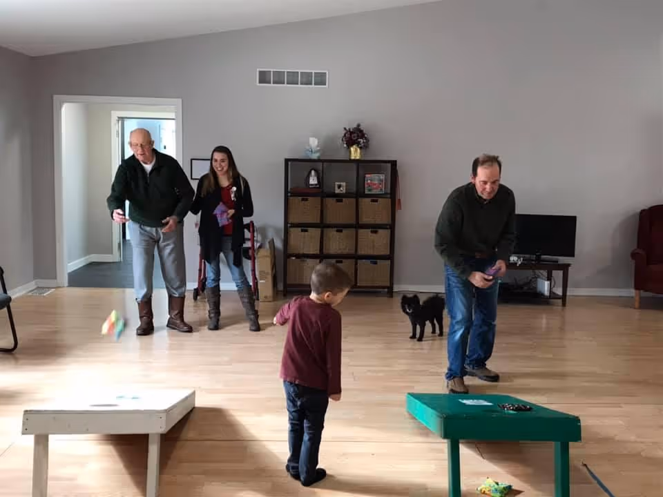 An elderly man, a middle-aged man, a young woman, and a young boy are playing a bean bag toss game in a spacious room with wooden floors. A small black dog is standing near the middle-aged man. The room has light gray walls, a shelving unit with baskets, a TV on a stand, and a red armchair in the corner.