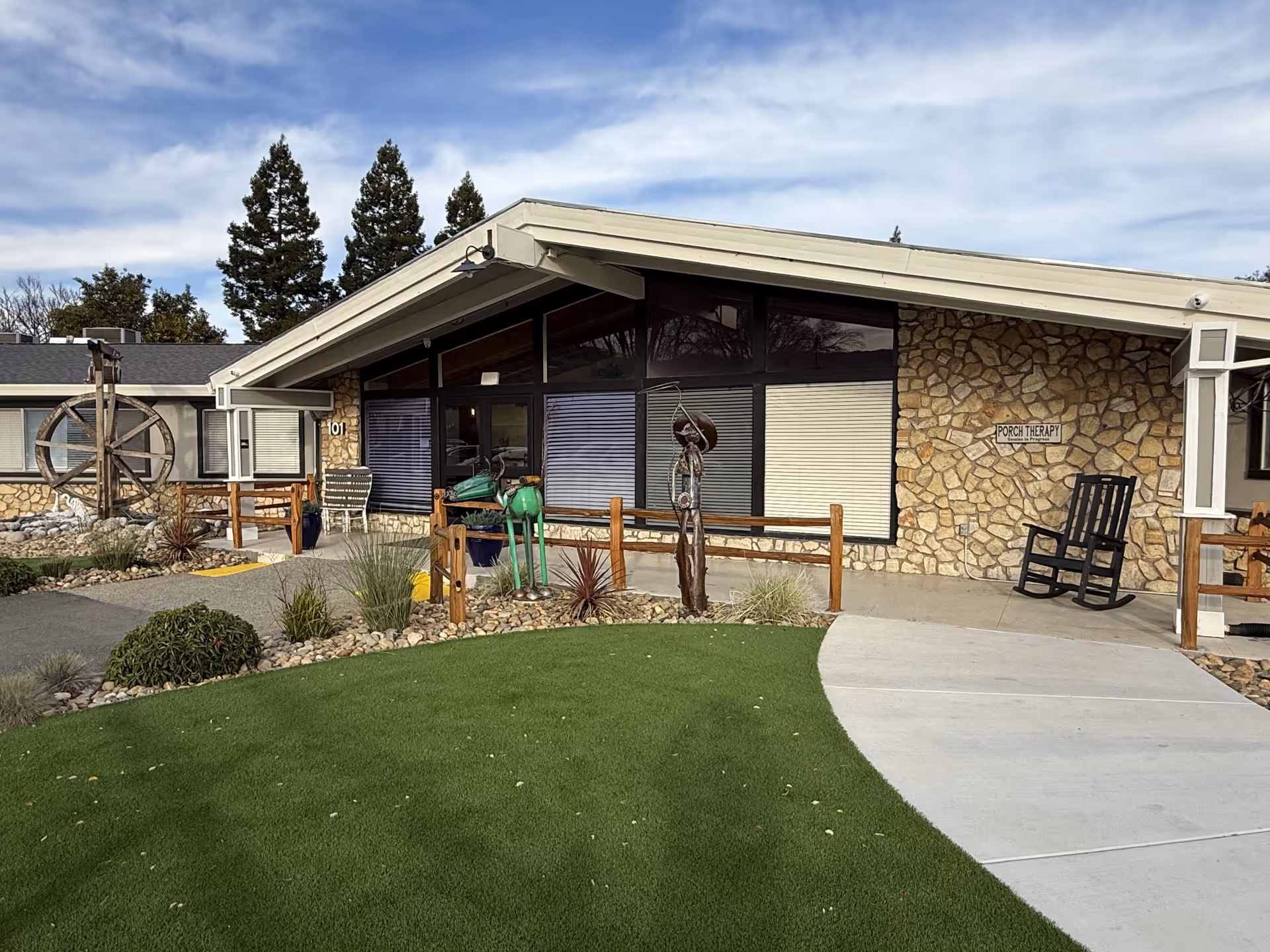 Exterior view of a single-story building with stone walls and large windows. There is a small garden with green grass and decorative plants in front. Two metal sculptures and a black rocking chair are placed near the entrance. A sign on the wall reads 'PORCH THERAPY'. The sky is partly cloudy with tall trees in the background.