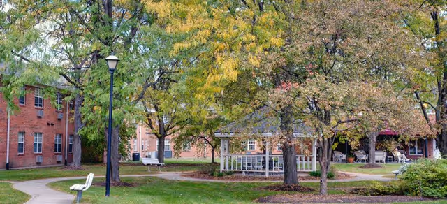 Outdoor view of a senior living facility with brick buildings, a gazebo, trees with autumn foliage, benches, and a lamppost along a winding paved pathway.