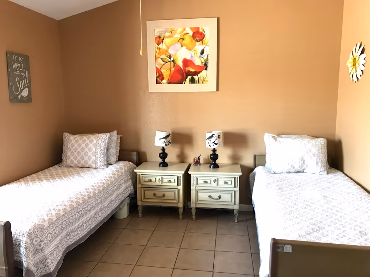 A bedroom with two single beds separated by two small white nightstands, each with a black lamp featuring a bird design. The walls are painted light brown and decorated with a colorful floral painting above the nightstands, a sign that reads 'It is well with my soul' on the left wall, and a large daisy decoration on the right wall. The floor is tiled.