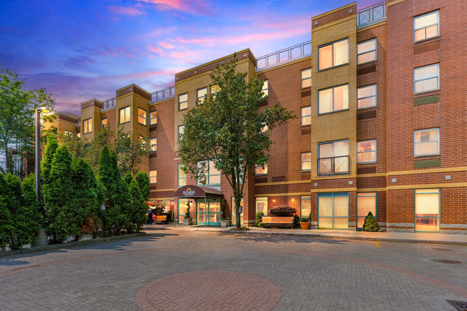 Exterior view of a multi-story brick building at sunset with a paved driveway and green trees. The entrance has a canopy with the sign 'Sunrise' above the door. The sky is colorful with shades of purple and pink.