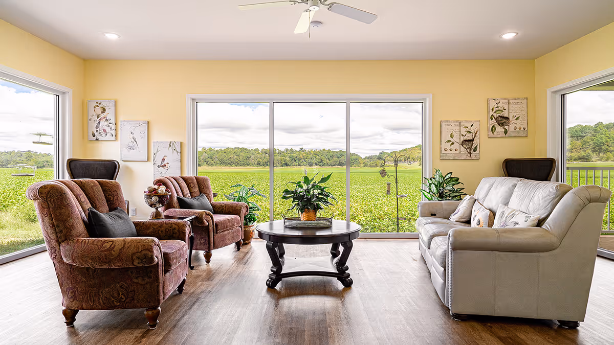 A bright living room with large windows offering a scenic view of green fields and trees. The room features two patterned armchairs with dark cushions on the left, a round wooden coffee table with a plant centerpiece in the middle, and a light-colored sofa with multiple cushions on the right. The walls are painted yellow and decorated with framed bird artwork. There are also several potted plants placed near the windows.