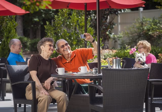 Two elderly people sitting at an outdoor table under a red umbrella, smiling and taking a selfie. Another elderly person is sitting in the background, and a woman holding a small white dog is seated at a nearby table. The setting is a garden or patio area with greenery and flowers.