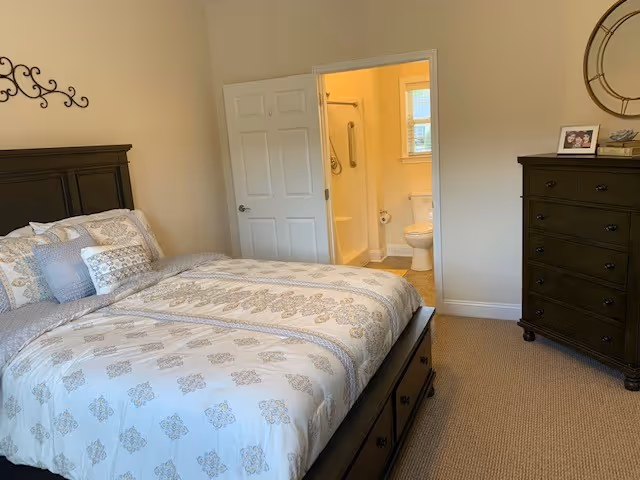 A bedroom with a large bed featuring patterned bedding and multiple pillows. To the right is a dark wooden dresser with a round wall clock above it and a framed photo on top. An open door reveals a bathroom with a toilet, grab bars, and a window.