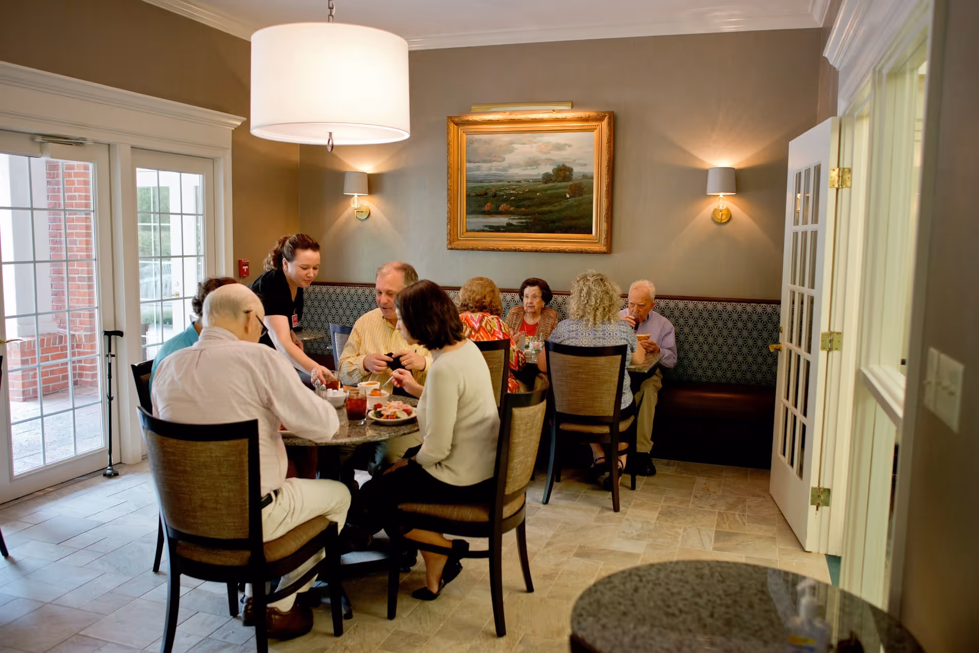 A group of elderly people sitting around a dining table in a well-lit room with a server attending to them. The room has beige walls, a large framed landscape painting, wall sconces, and a large hanging light fixture. There are glass doors leading outside and a patterned bench along one wall.