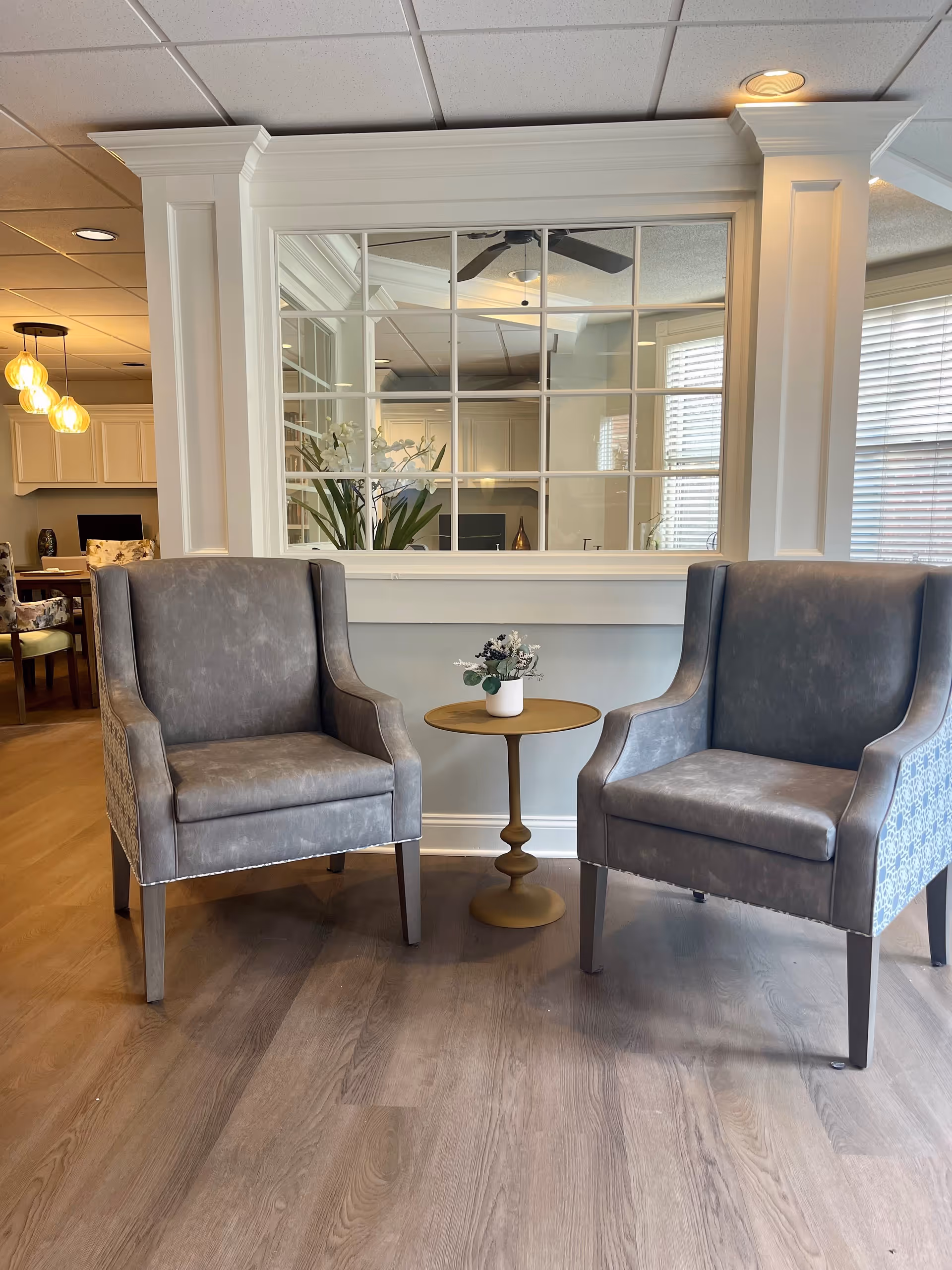 Two gray upholstered armchairs with patterned sides are positioned on either side of a small round wooden table with a small white vase containing flowers. Behind the chairs is a large window with multiple panes, reflecting the interior of a room with ceiling lights and a ceiling fan. The floor is wood, and the walls are light-colored with white trim.