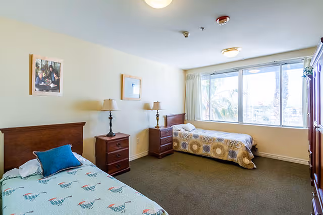 A bright bedroom with two single beds, each with a wooden headboard and bedside table with a lamp. The room has a large window letting in natural light and a wooden wardrobe on the right side. There are two framed pictures on the wall above the beds.