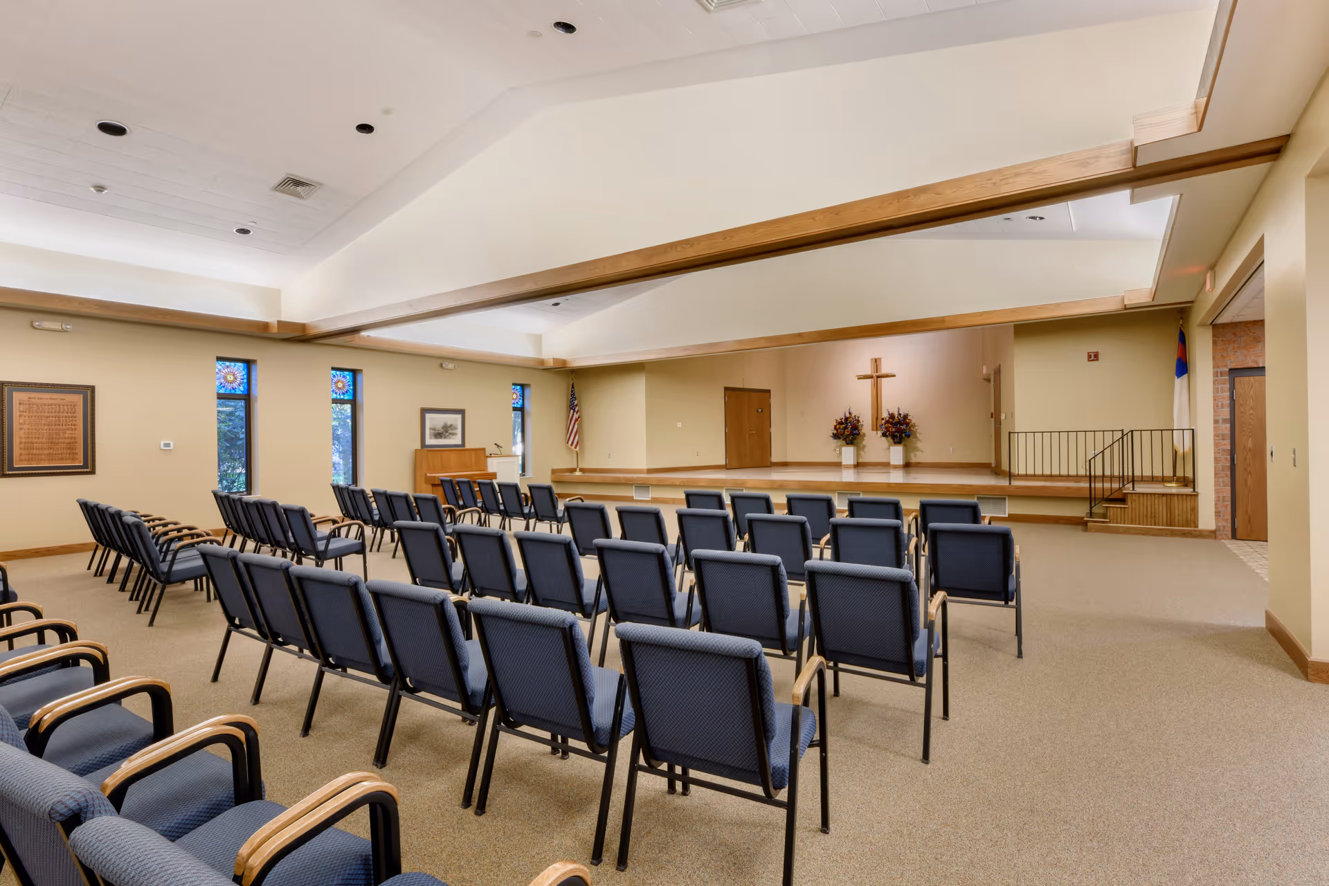Interior view of a chapel or meeting room with rows of blue cushioned chairs facing a raised stage. The stage has a wooden cross on the wall, two flower arrangements, and an American flag to the left. The room has beige walls, a high ceiling with wooden beams, and several narrow stained glass windows along one wall.