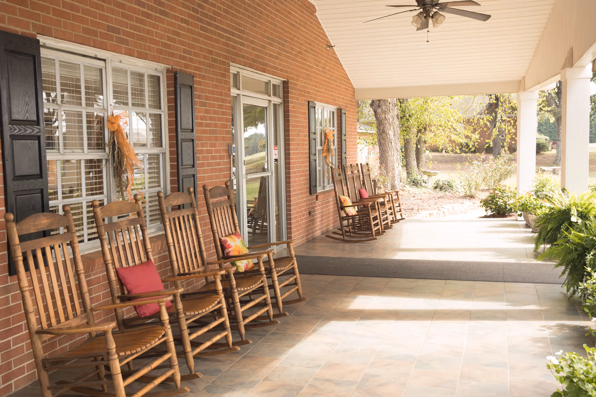 Covered outdoor porch area with a row of wooden rocking chairs, some with colorful cushions, against a brick wall with windows and a glass door. The porch overlooks a garden area with trees and plants, and there is a ceiling fan above.