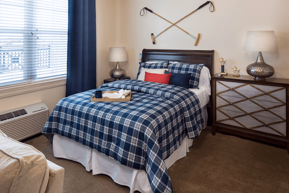 A cozy bedroom with a bed dressed in blue and white plaid bedding, accented with red and navy pillows. Two crossed golf clubs are mounted on the wall above the dark wooden headboard. There is a nightstand on each side of the bed, each with a lamp. A window with white blinds and navy curtains is on the left side, and a beige armchair is partially visible in the foreground.
