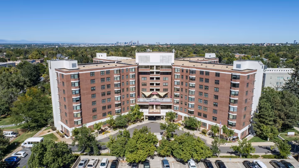 Aerial frontal view of a multi-story red-brick senior living building with a circular driveway, trees, and parked cars.