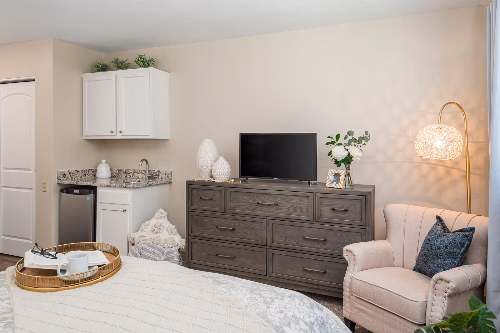 A cozy bedroom corner featuring a beige armchair with a blue patterned pillow, a wooden dresser with a flat-screen TV, decorative vases, a framed photo, and a vase with white flowers. To the left, there is a small kitchenette area with a granite countertop, a sink, white cabinets, and a mini fridge. A tray with a book, glasses, and a coffee cup is placed on the bed in the foreground.
