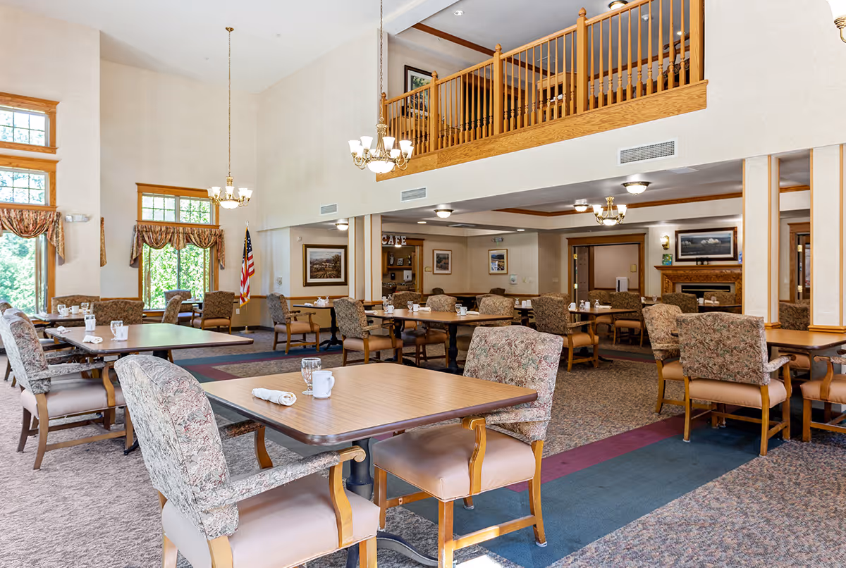 A spacious dining room with multiple wooden tables and cushioned chairs arranged neatly. The room features large windows with patterned curtains allowing natural light to enter. There are chandeliers hanging from the ceiling, an American flag in the corner, and a second-floor balcony with wooden railings overlooking the area. The walls are decorated with framed artwork and there is a fireplace in the background.