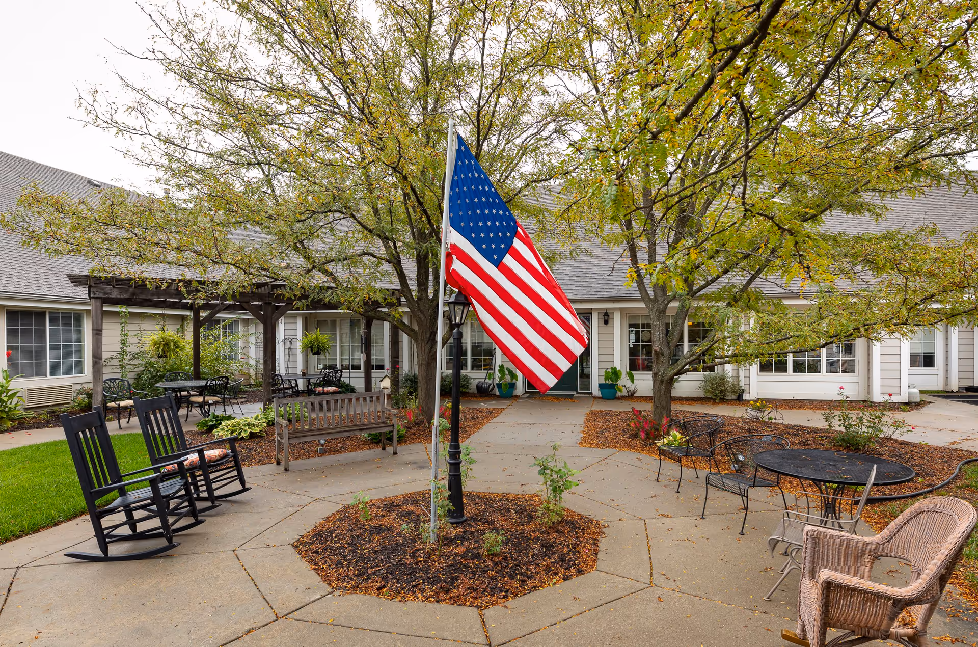 Outdoor courtyard area at Homestead Assisted Living of Leavenworth featuring an American flag on a pole in the center, surrounded by trees, benches, rocking chairs, and patio tables with chairs. The building exterior with windows and doors is visible in the background.