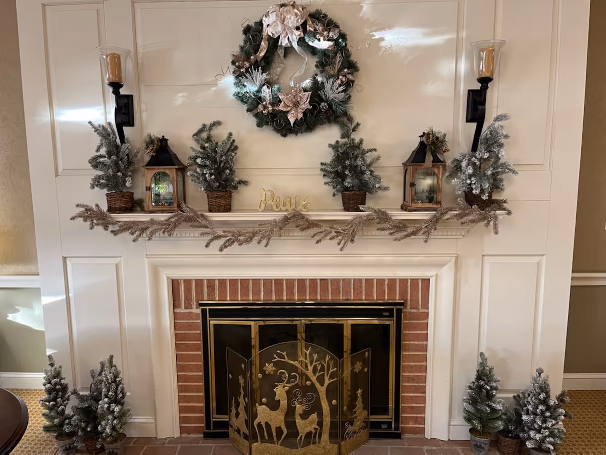Fireplace mantel decorated for winter with a wreath, lanterns, frosted miniature trees, garland, and a deer-themed fireplace screen.
