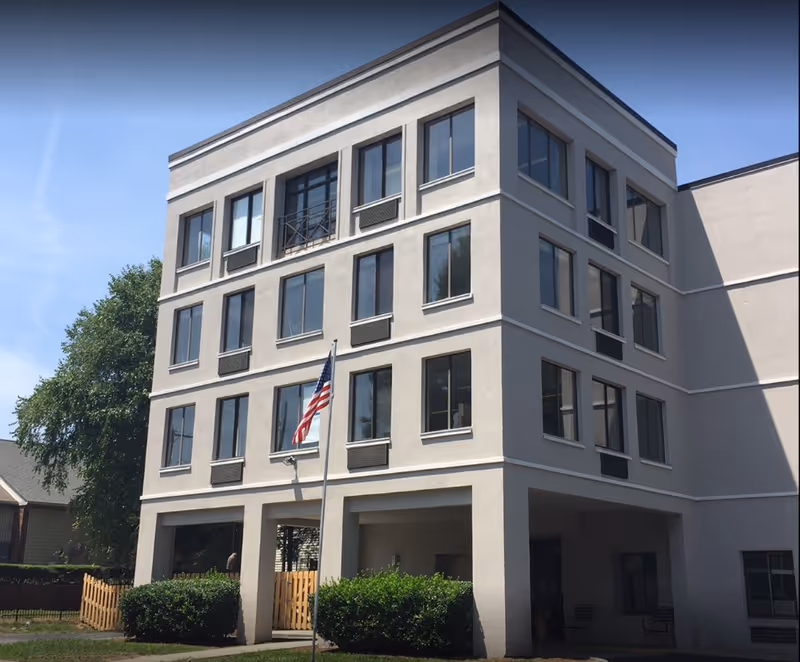 Exterior view of a four-story beige building with multiple windows, an American flag on a pole in front, and green bushes near the entrance. The sky is clear and blue.