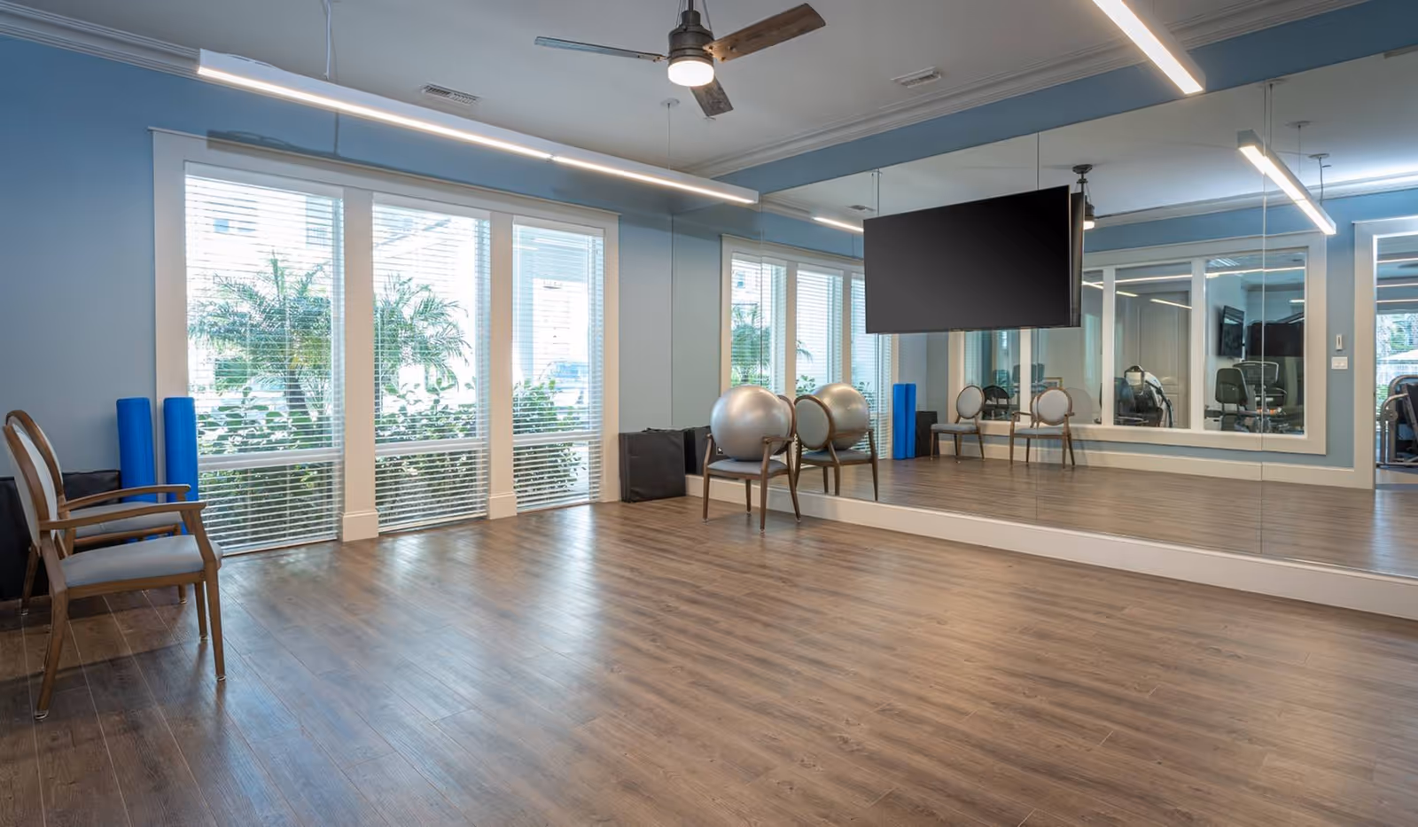 A bright exercise room with wooden flooring, light blue walls, large windows with white blinds, a ceiling fan, a wall-mounted TV, and a large mirror covering one wall. There are two chairs with exercise balls on them and two blue foam rollers leaning against the wall.