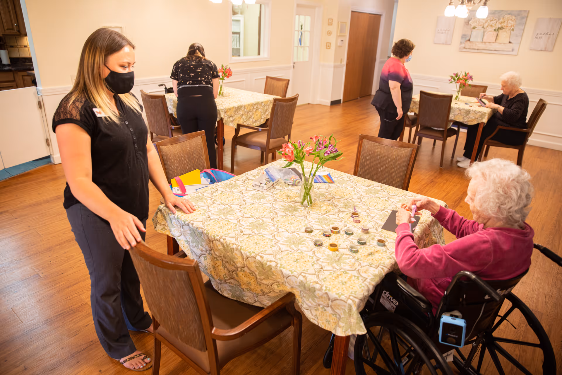 Staff and residents wearing masks gather around floral-covered tables in a senior living dining/activity room while an elderly woman in a wheelchair works on a craft.