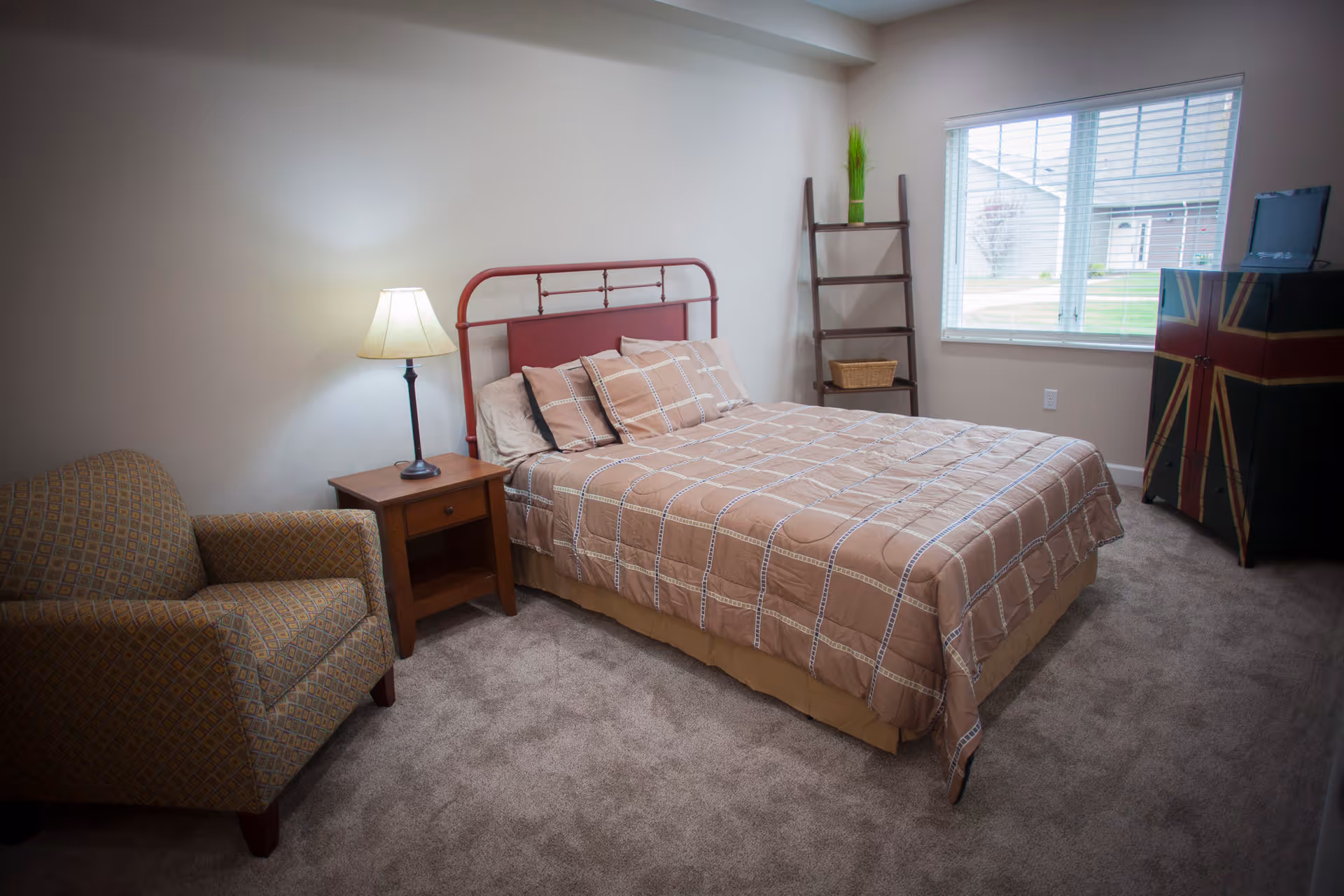 A cozy bedroom with a double bed featuring a red metal headboard and beige checkered bedding. Next to the bed is a wooden nightstand with a lamp. There is a patterned armchair to the left and a wooden ladder shelf with a green plant and basket near the window. A cabinet with a Union Jack design is positioned under the window, which shows a view of the outside buildings.