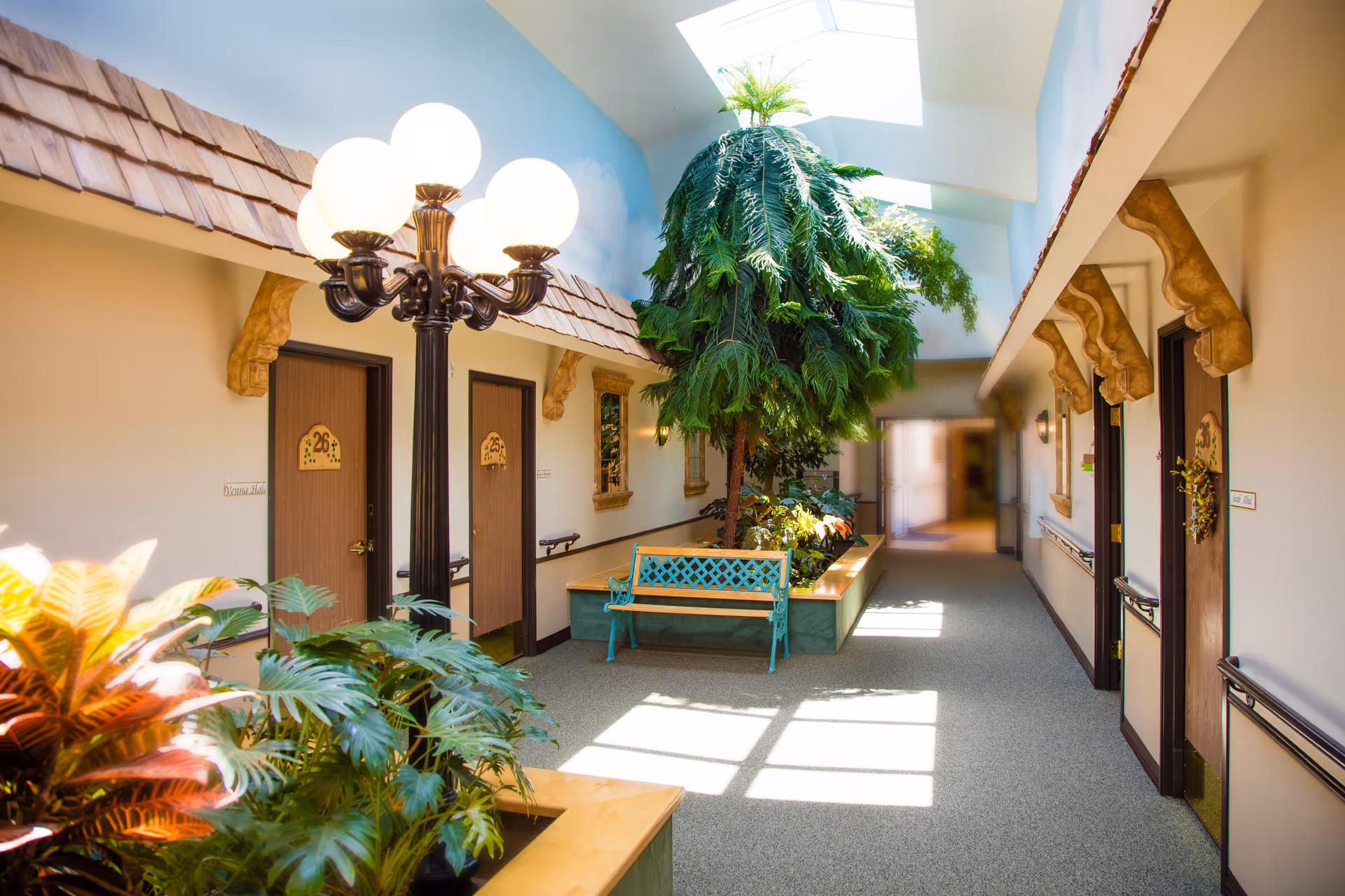 Bright indoor hallway in a senior living facility with wooden doors numbered 25, 26, and 36, decorative wooden awnings above the doors, a central planter with a large green tree and other plants, a turquoise bench, and a vintage-style lamp post with five glowing globes. Skylights allow natural light to illuminate the space.