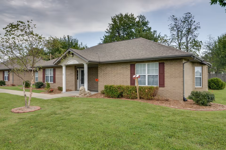 Exterior view of a single-story brick building with a gray shingled roof, white columns at the entrance, and red shutters on the windows. The building is surrounded by green grass, small trees, and shrubs under a cloudy sky.