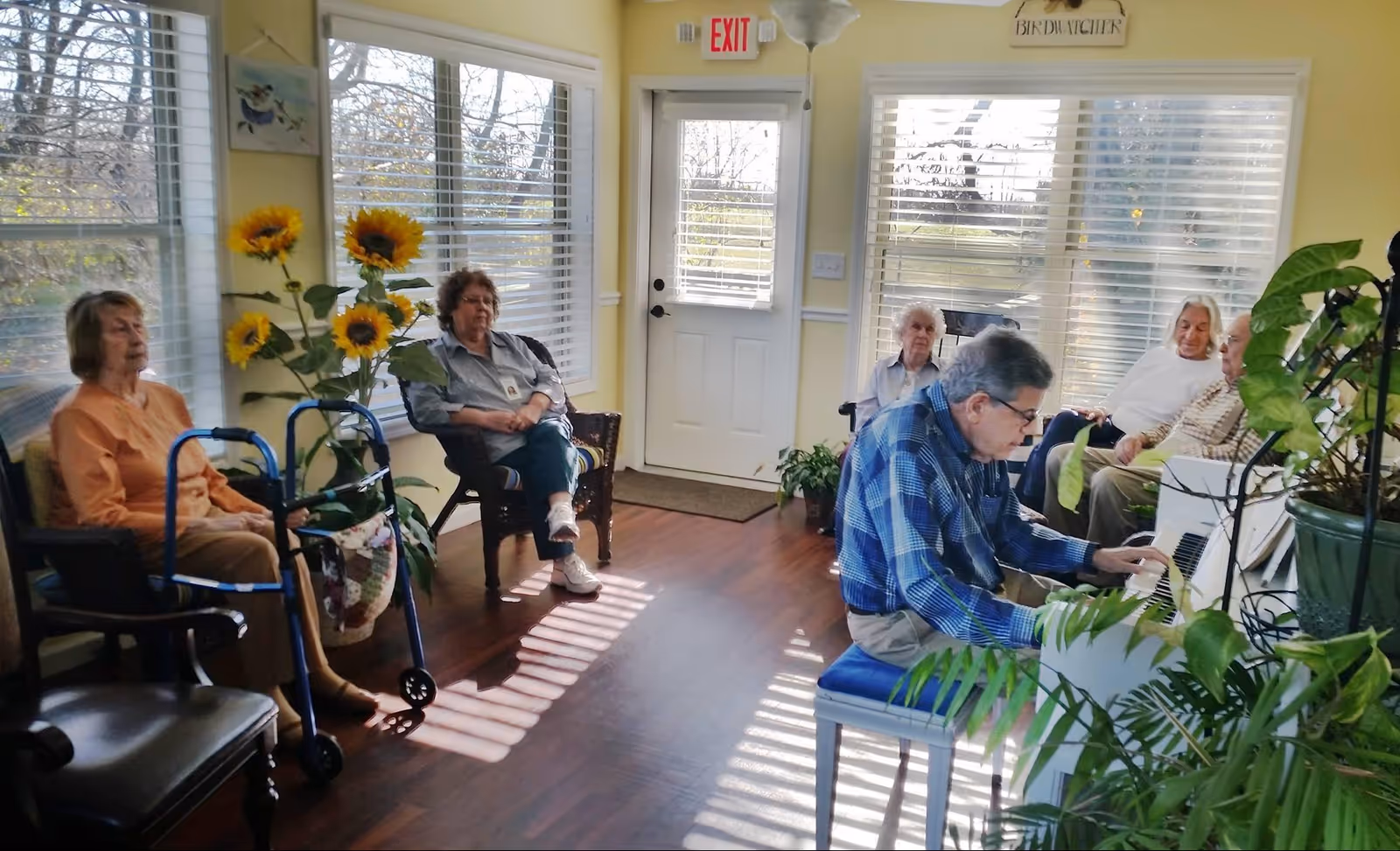 A group of elderly people sitting in a bright room with large windows and wooden floors. One man is playing a white piano while others sit on chairs around the room, listening. There are plants and sunflowers decorating the space, and an exit door is visible in the background.