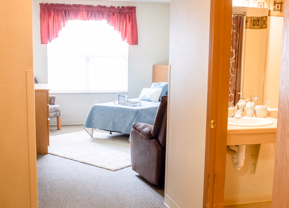 View into a senior living bedroom with a bed covered in a blue bedspread, a brown recliner chair, a dresser, and a window with red curtains. To the right, there is a partial view of a bathroom sink and mirror.