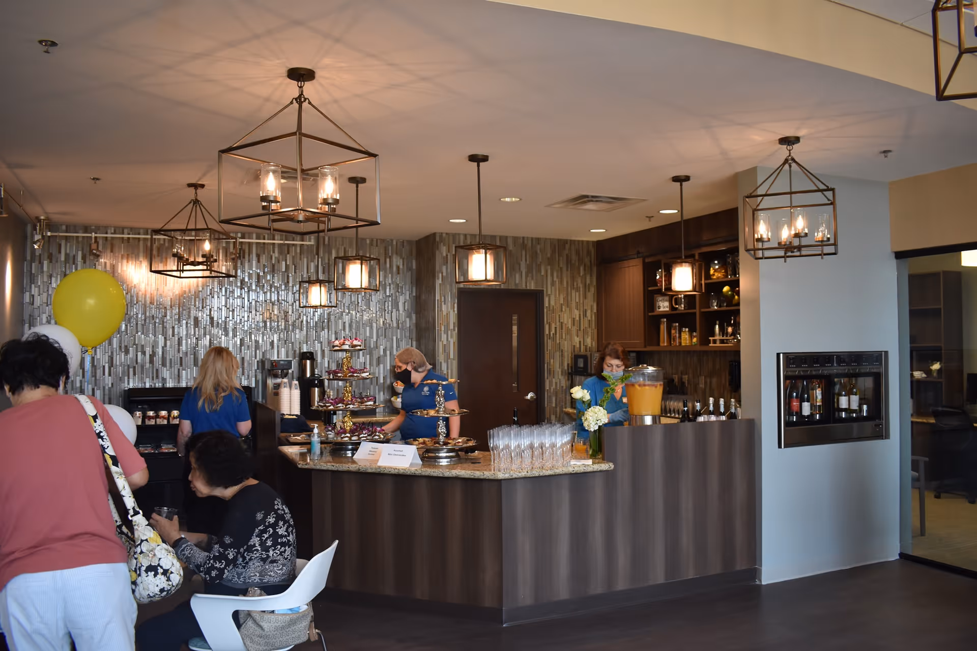 Interior view of a senior living facility's dining or refreshment area with a counter displaying drinks, glasses, and snacks. Several people are present, some seated and some standing behind the counter serving. The room features modern pendant lighting and a tiled wall backdrop.