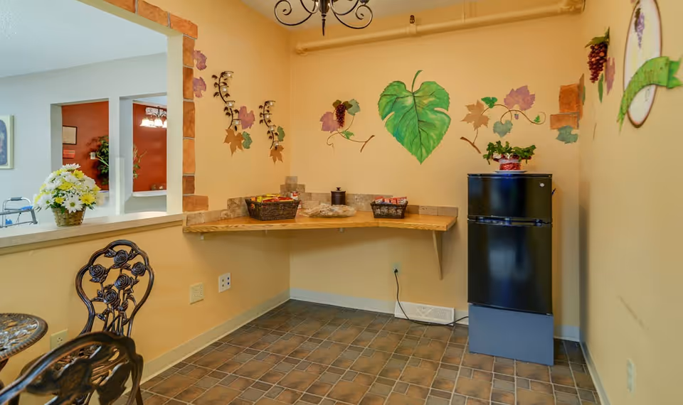 A small corner area in a senior living facility with a black mini refrigerator, a wooden corner shelf holding baskets and snacks, and walls decorated with painted grapevine murals. There is a small round metal table with chairs nearby and a window opening to another room with a walker and flowers visible.