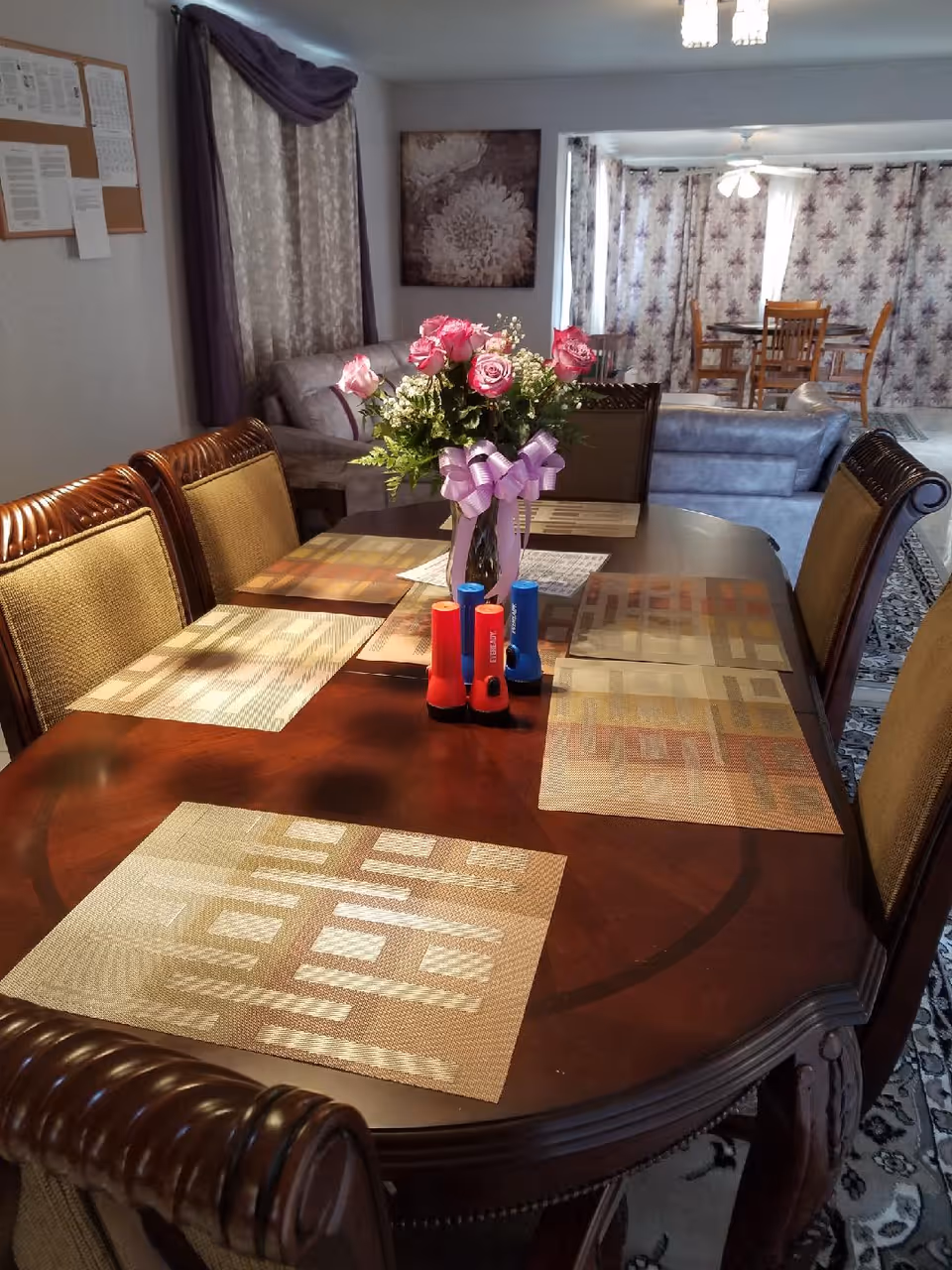 A dining room with a dark wooden table set with six placemats and a vase of pink roses with a purple ribbon in the center. There are four chairs visible around the table. In the background, there is a living area with a sofa, a wall painting, and another dining table with chairs near windows covered by floral curtains.
