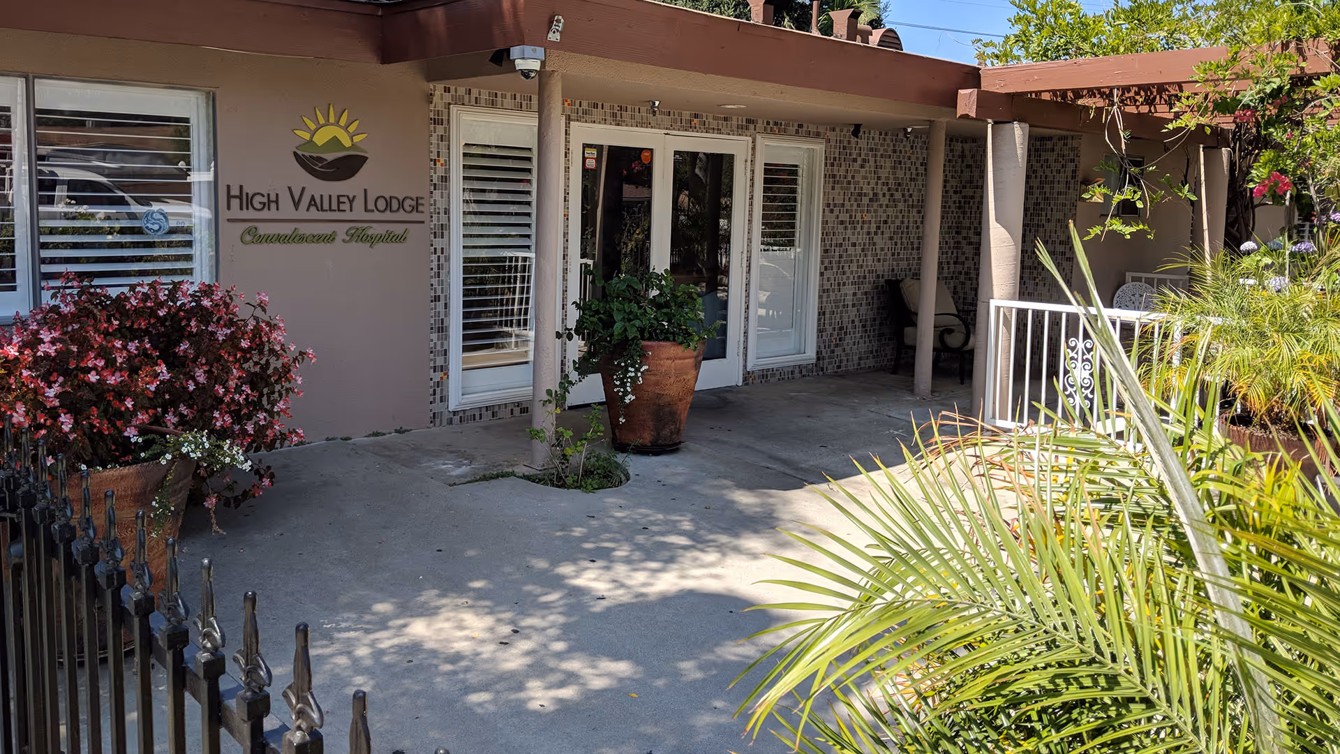 Entrance of High Valley Lodge Convalescent Hospital with a covered porch, potted plants, and a decorative fence. The building exterior features a tiled wall section and windows with white shutters.